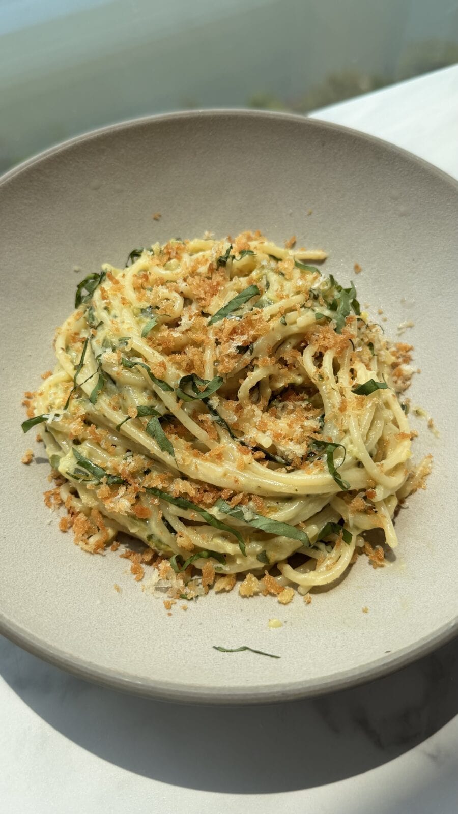 A bowl of spaghetti pasta topped with shredded greens and crispy breadcrumbs, served in a neutral-colored bowl on a light surface.