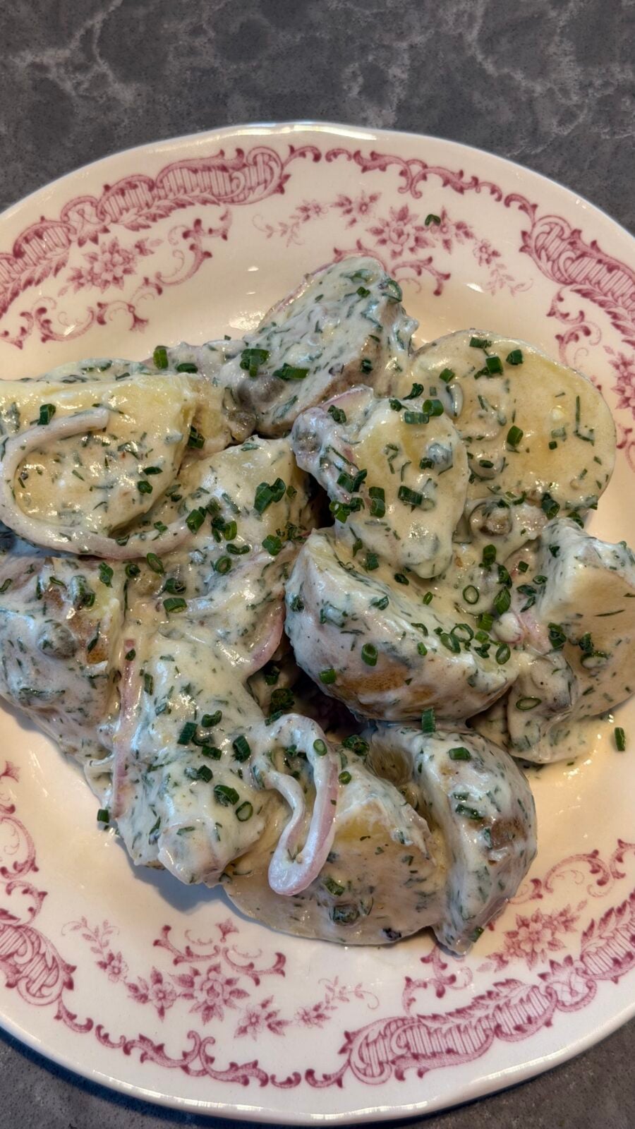 Potato salad with creamy herb dressing and sliced onions, served on a decorative plate with a pink floral border, on a gray countertop.