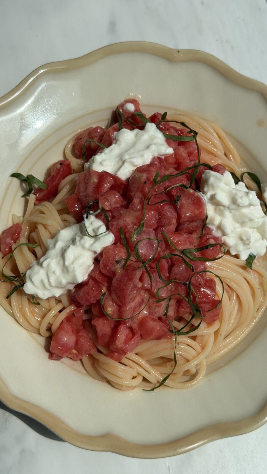 A plate of spaghetti topped with chopped tomatoes, fresh basil, and dollops of ricotta or burrata cheese on a white dish.