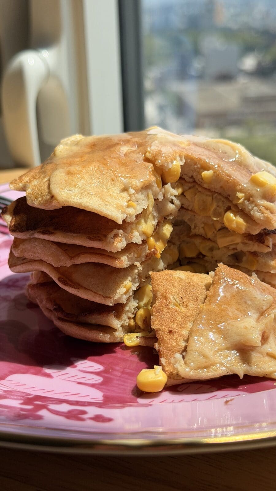 A stack of pancakes with corn kernels inside, cut to show the filling, sits on a pink plate by a window in natural light.
