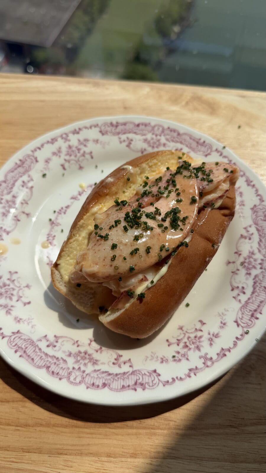 A lobster roll in a toasted bun topped with sauce and chopped herbs, served on a decorative white plate with a pink floral border.