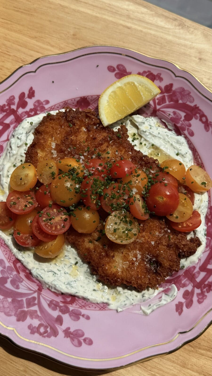 Breaded cutlet on creamy herb sauce, topped with halved cherry tomatoes and chopped chives, served with a lemon wedge on a pink floral plate.