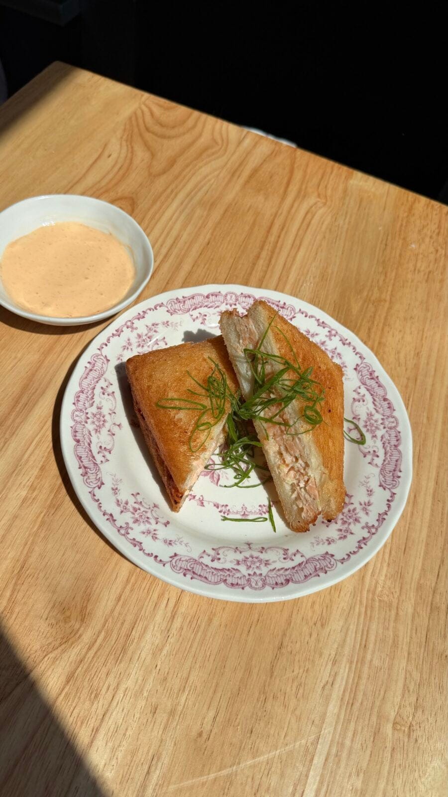 A toasted sandwich cut in half with green garnish on a patterned plate, served with a small bowl of dipping sauce on a wooden table.
