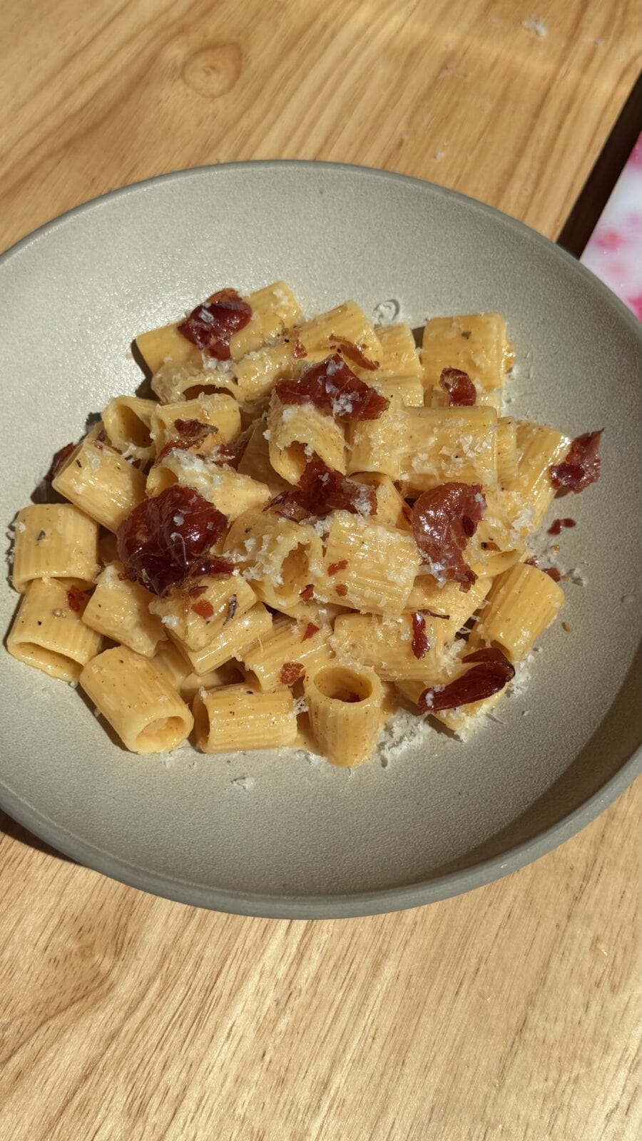 A plate of rigatoni pasta topped with grated cheese and pieces of cured meat, served on a light-colored dish on a wooden table.
