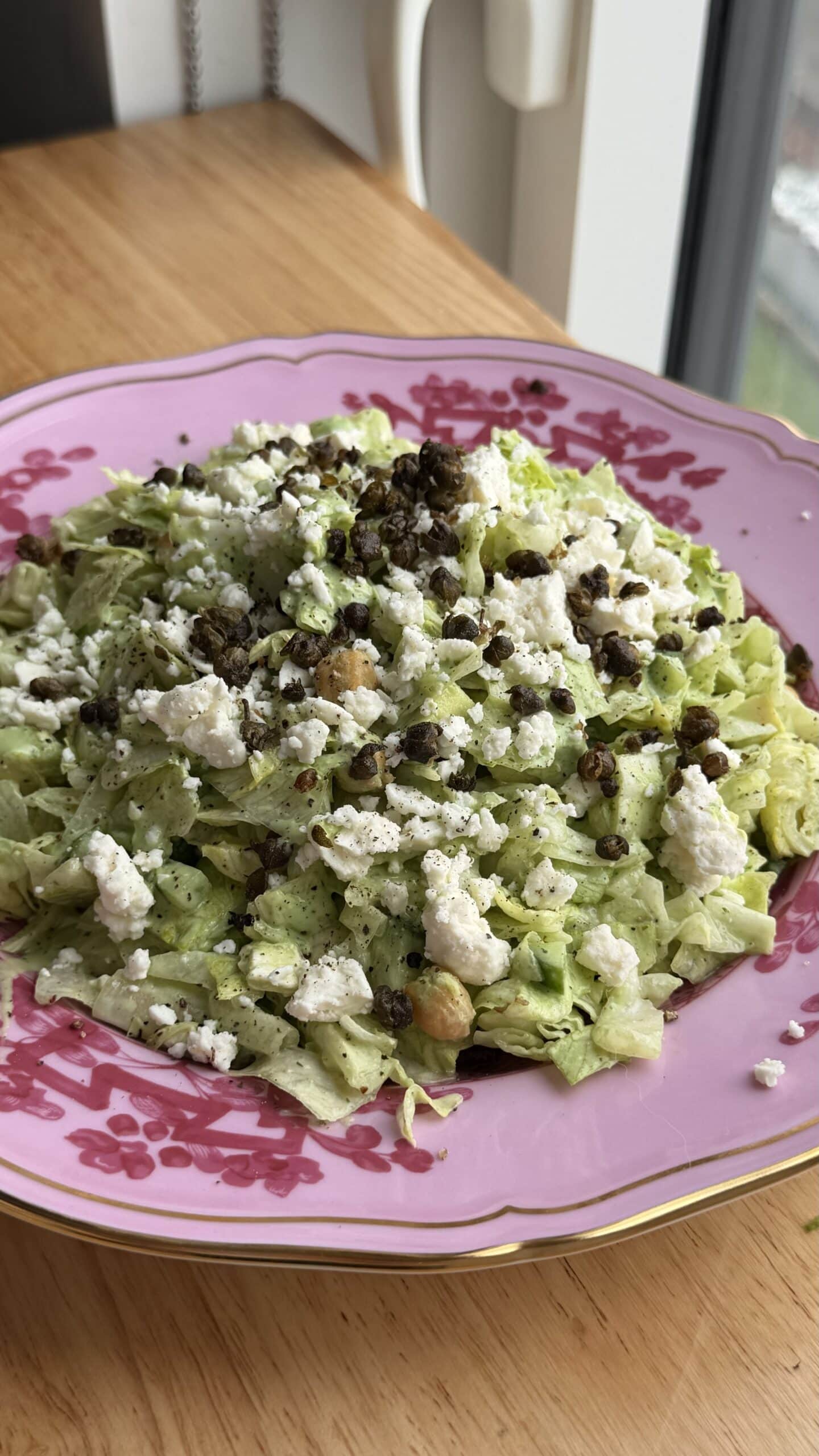 A plate of chopped lettuce salad topped with crumbled cheese, black lentils, and chickpeas, served on a pink floral dish by a window.
