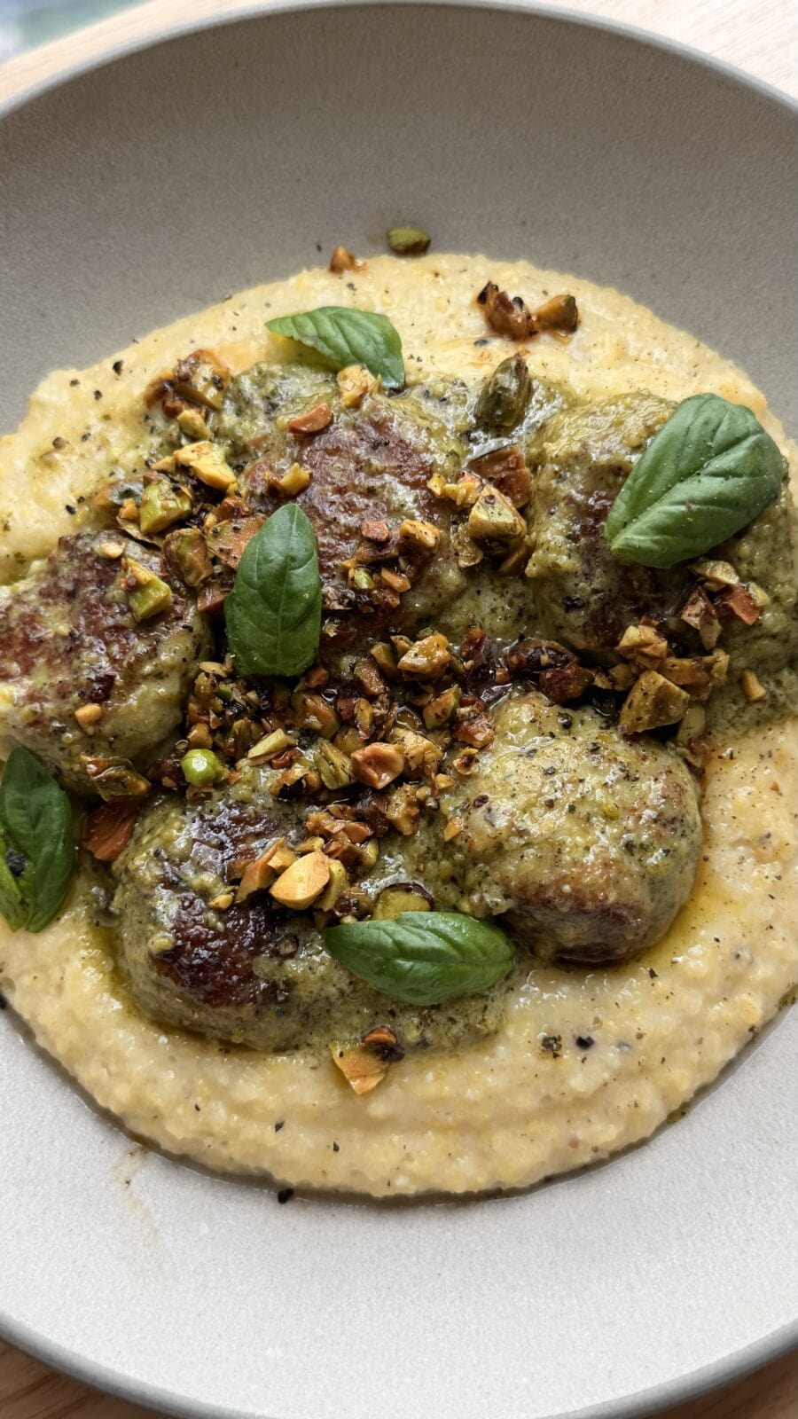 Meatballs on creamy polenta, topped with pesto, chopped pistachios, black pepper, and fresh basil leaves, served in a beige bowl.