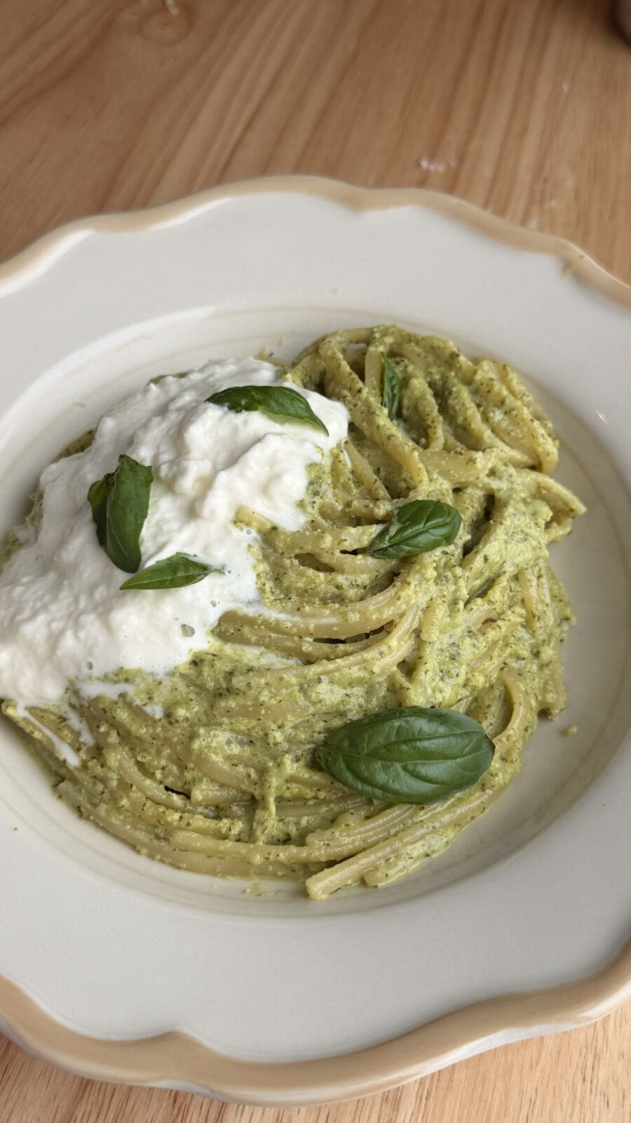 A plate of pasta with green pesto sauce, topped with a dollop of white cheese and garnished with fresh basil leaves on a wooden table.