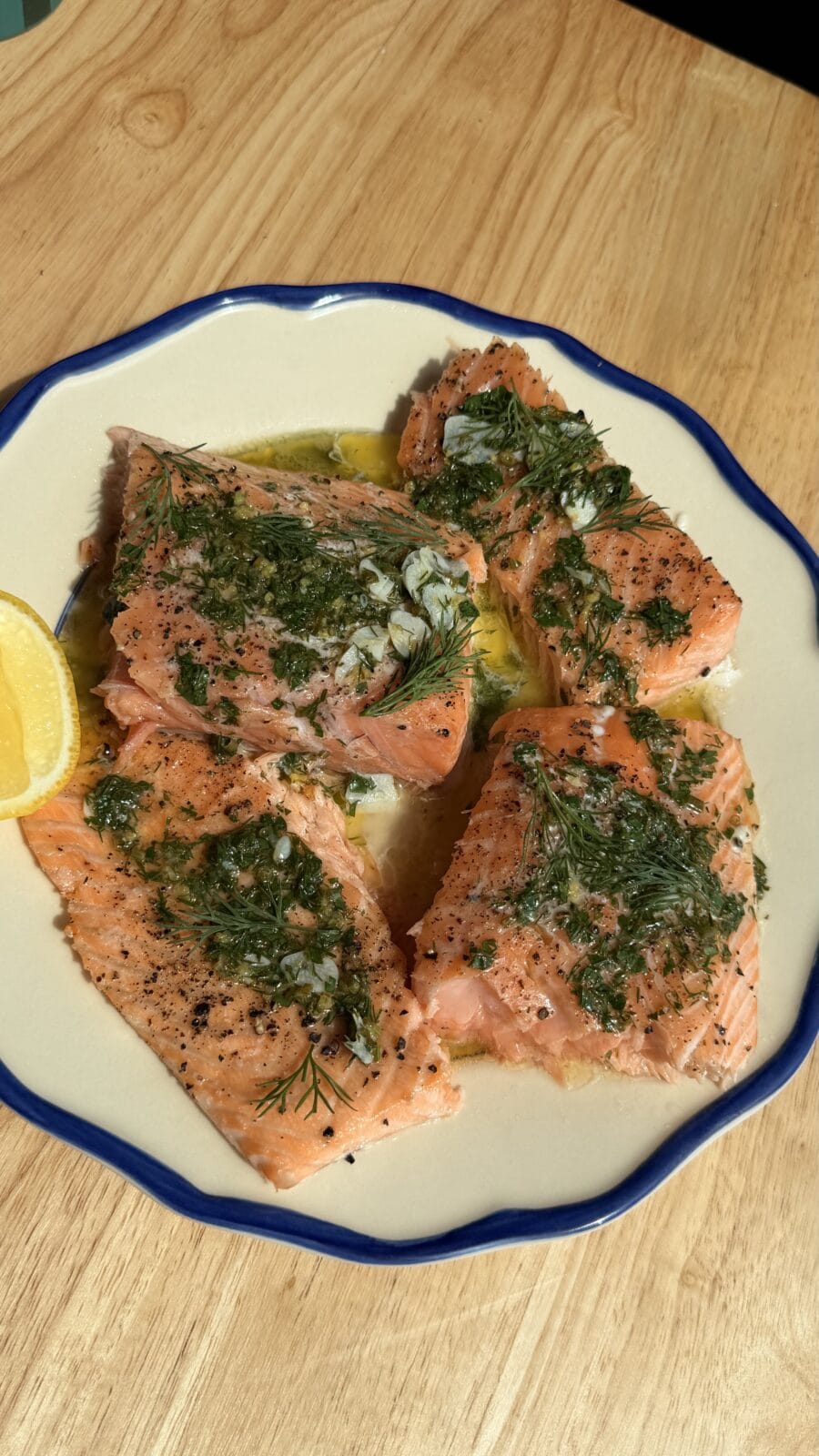 Four salmon fillets seasoned with herbs and garlic on a plate with a lemon wedge.