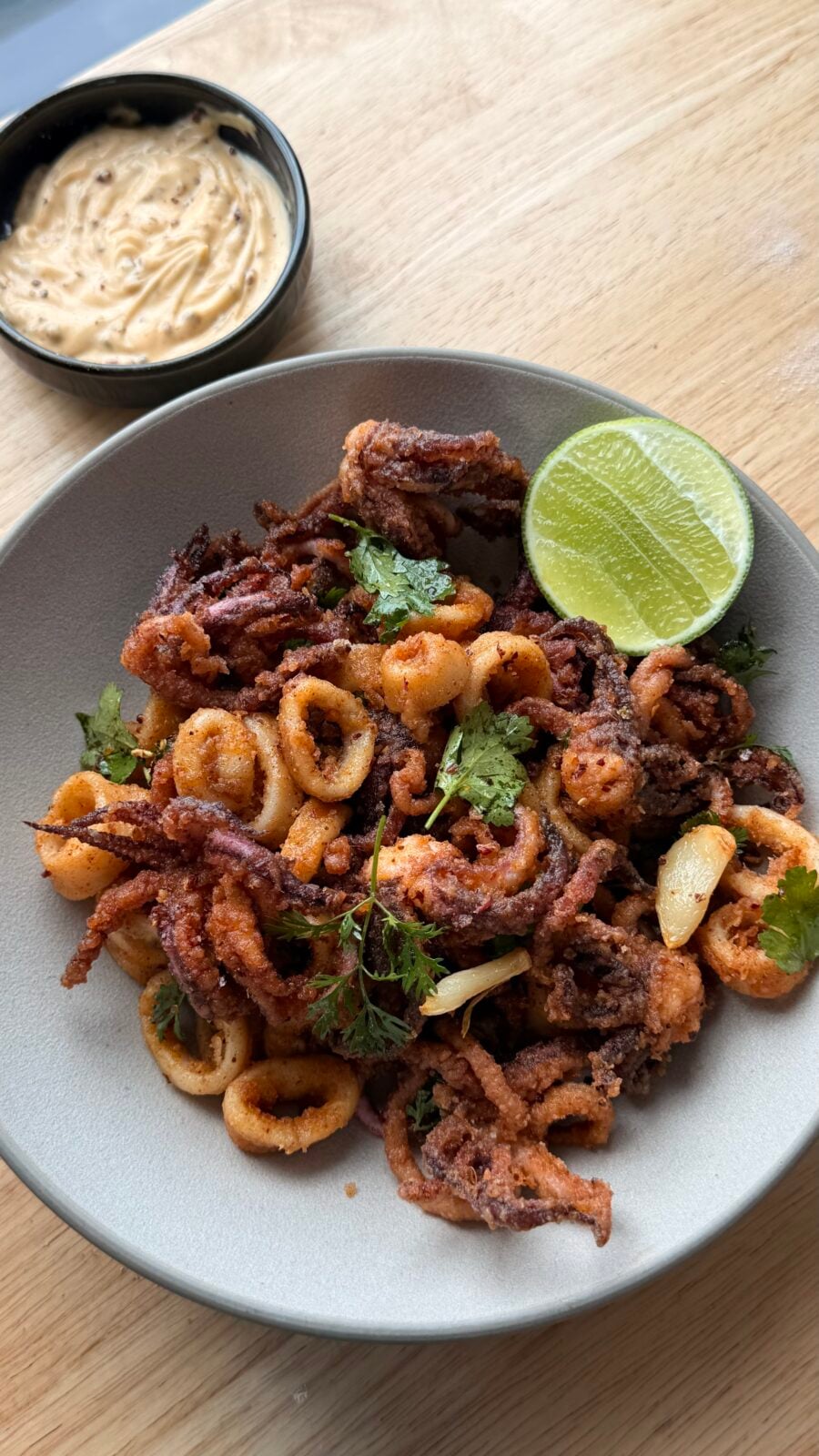 A bowl of fried calamari garnished with cilantro and a lime wedge, served with a small dish of creamy dipping sauce on a wooden surface.