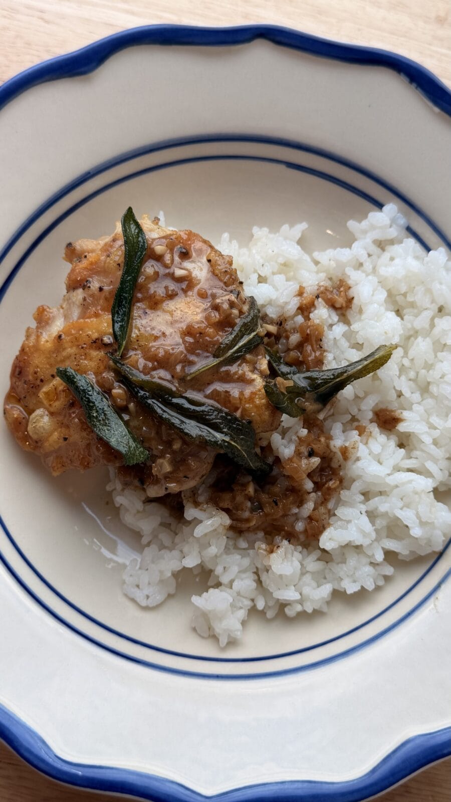 A piece of chicken with brown sauce and herbs served over white rice on a round plate with blue trim.