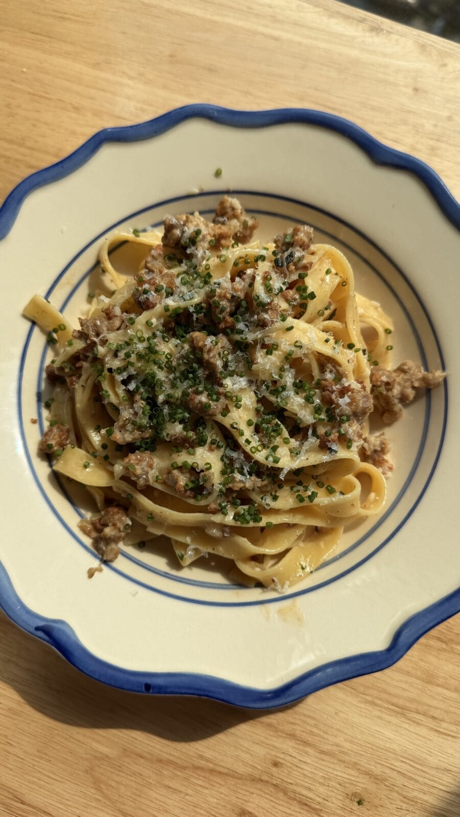 A plate of tagliatelle pasta topped with ground meat, grated cheese, and chopped herbs, served on a white dish with blue trim.