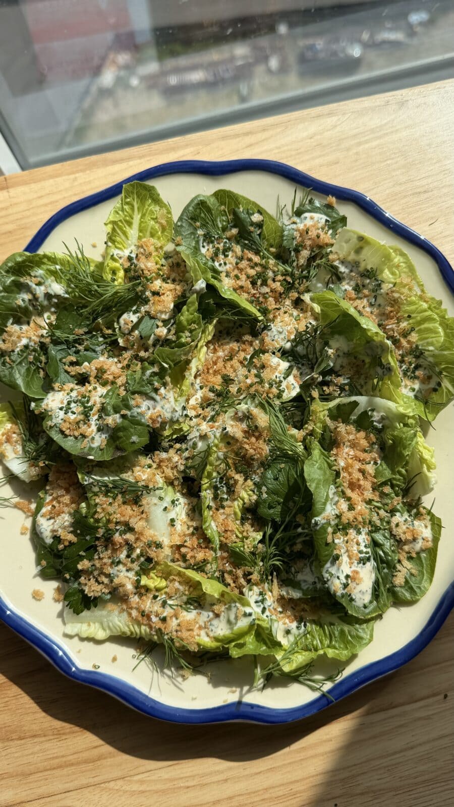 A plate of leafy green salad topped with breadcrumbs, fresh herbs, and a creamy dressing, placed on a wooden surface in sunlight.