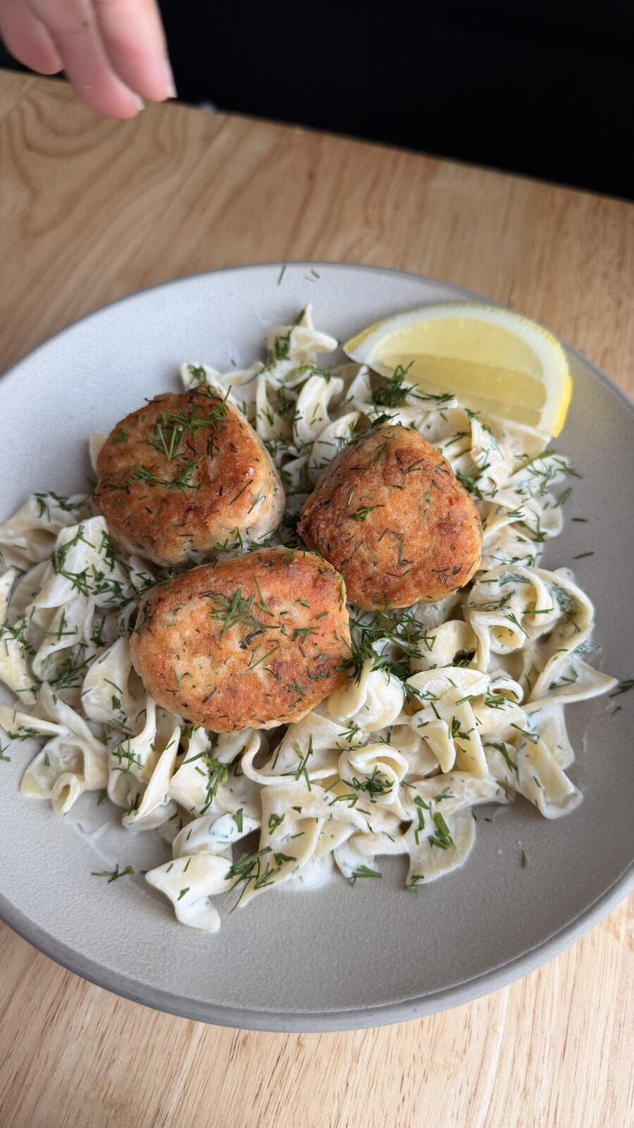 Three golden brown fish cakes on a bed of pasta with creamy sauce, sprinkled with herbs, and served with two lemon wedges on a gray plate.