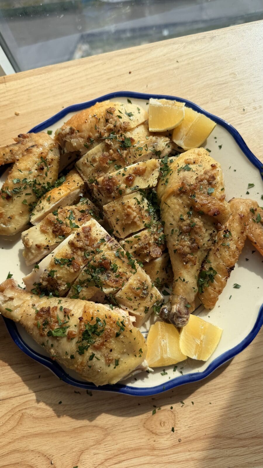 A plate of roasted chicken pieces garnished with herbs, accompanied by lemon wedges, on a wooden surface in natural sunlight.