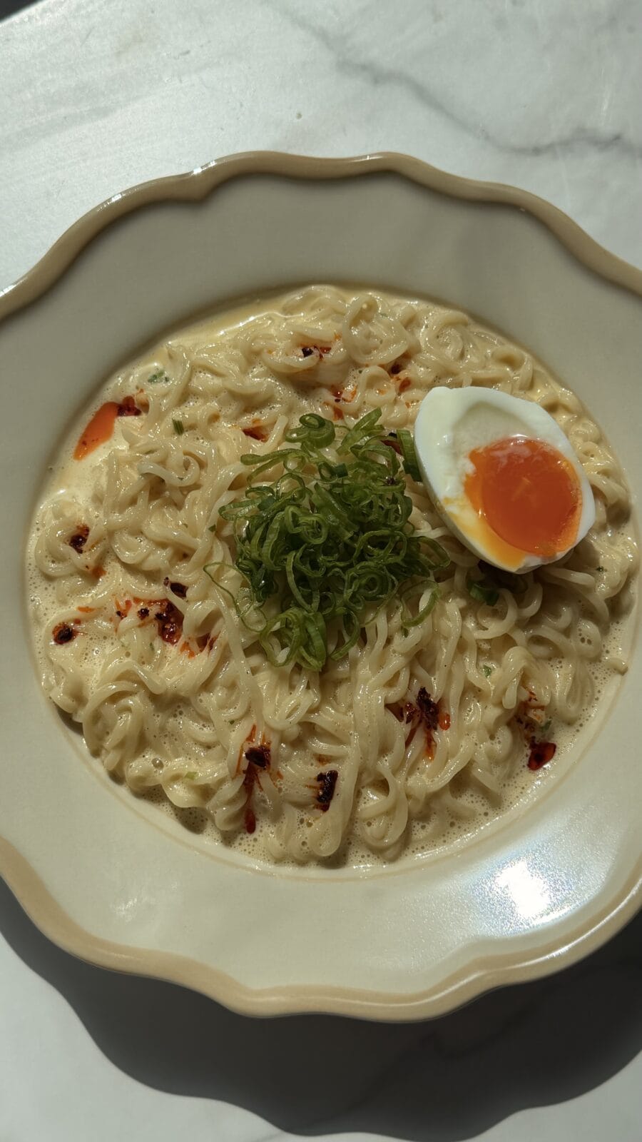 Bowl of ramen noodles in creamy broth, topped with sliced green onions, chili flakes, and half of a soft-boiled egg.