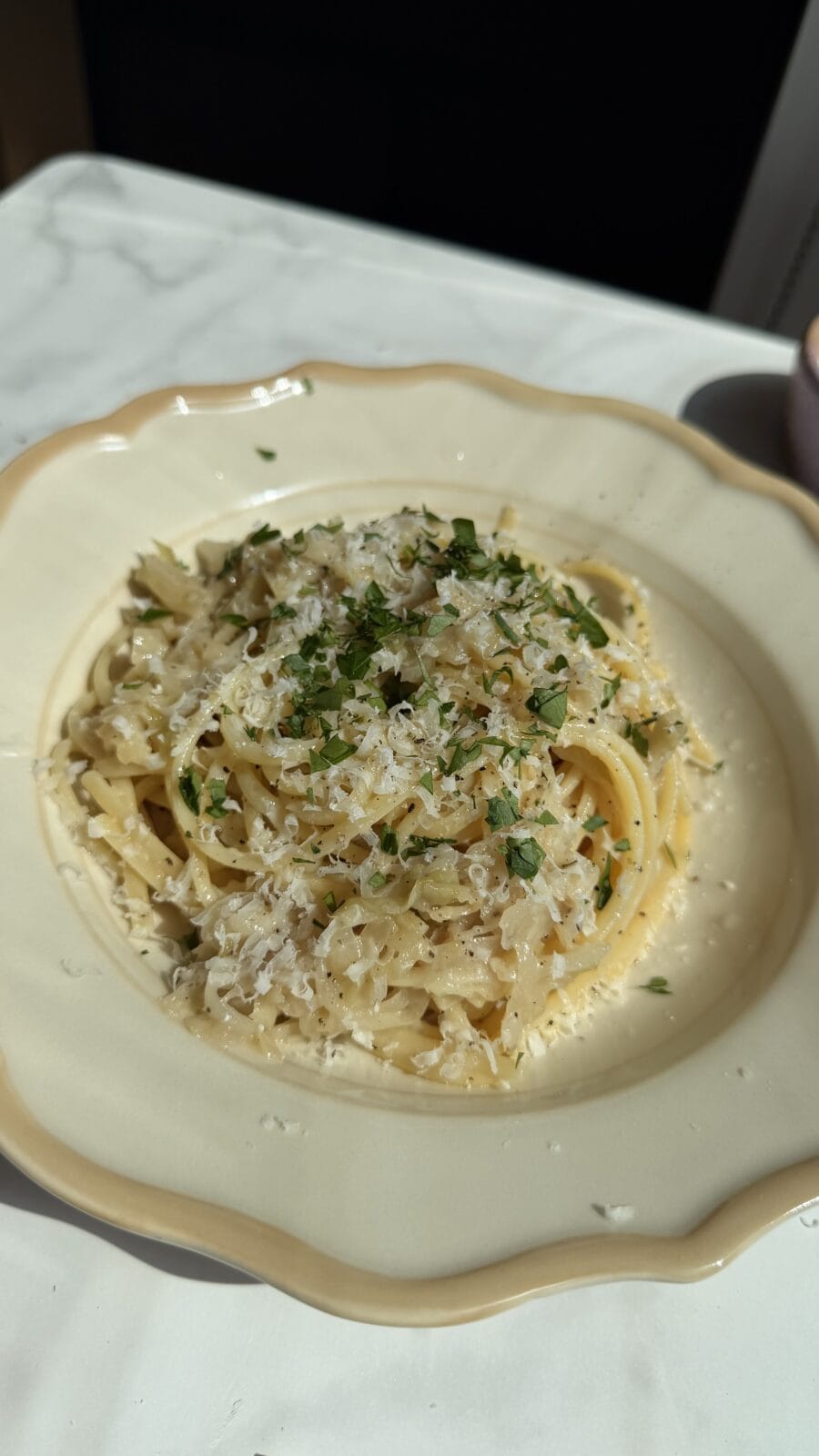 A plate of spaghetti topped with grated cheese and chopped herbs, served on a cream-colored dish on a white marble surface.