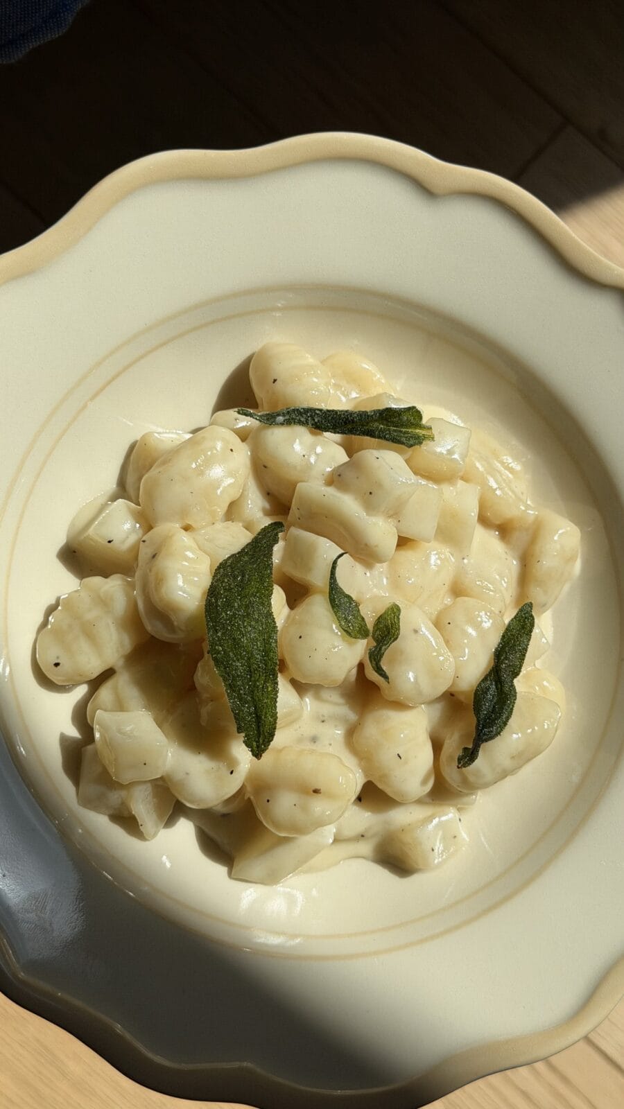 A plate of gnocchi in a creamy white sauce, garnished with crispy sage leaves, served in a cream-colored bowl.