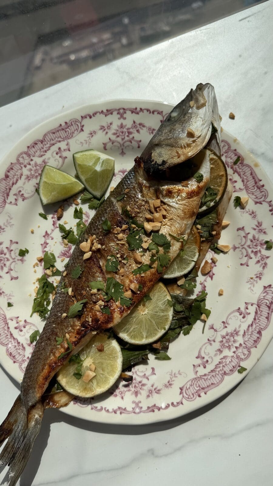 A whole roasted fish garnished with chopped herbs and nuts, served on a plate with lime slices and lime wedges.