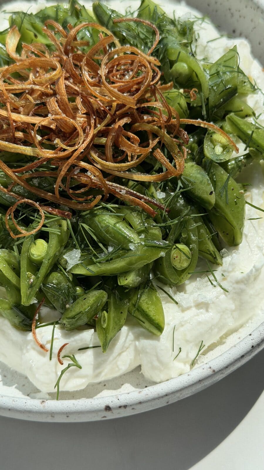 A plate of whipped ricotta topped with sliced snap peas, fresh herbs, and crispy fried shallots.
