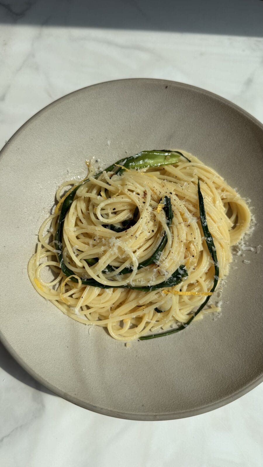A plate of spaghetti topped with grated cheese and green vegetables, served on a light-colored dish against a marble background.