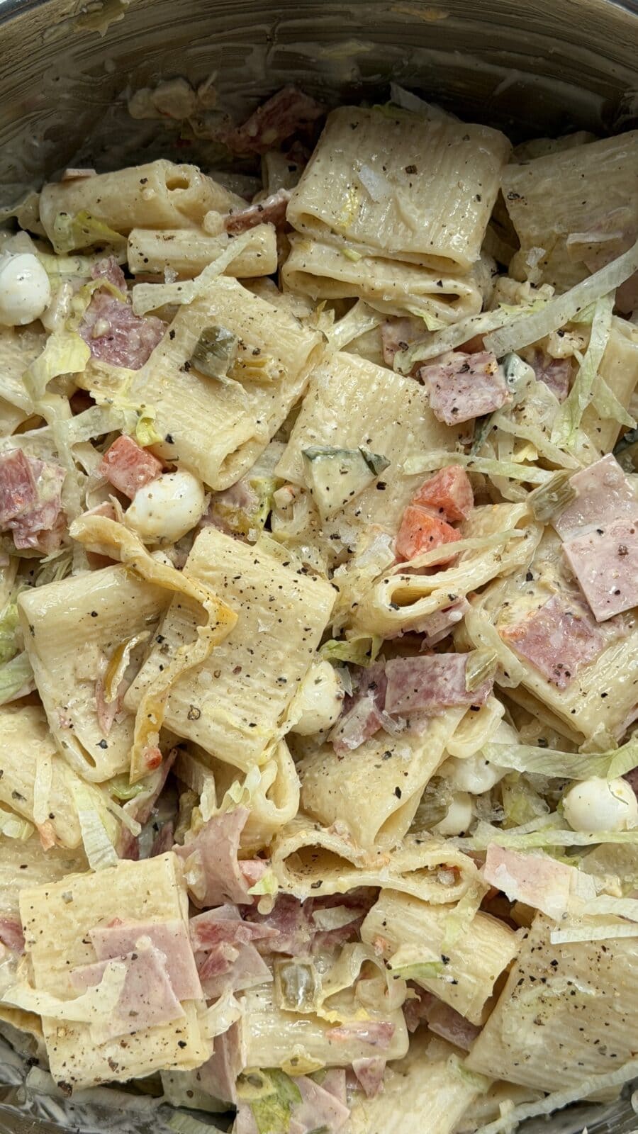 A close-up of pasta salad with rigatoni, diced deli meats, mozzarella balls, lettuce, pickles, tomatoes, and black pepper in a creamy dressing.