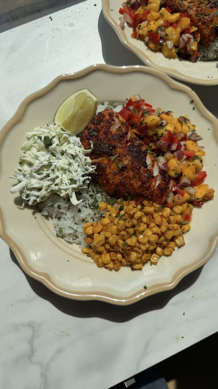 A plate with blackened fish, mango salsa, grilled corn, shredded cabbage, a lime wedge, and rice on a beige dish.