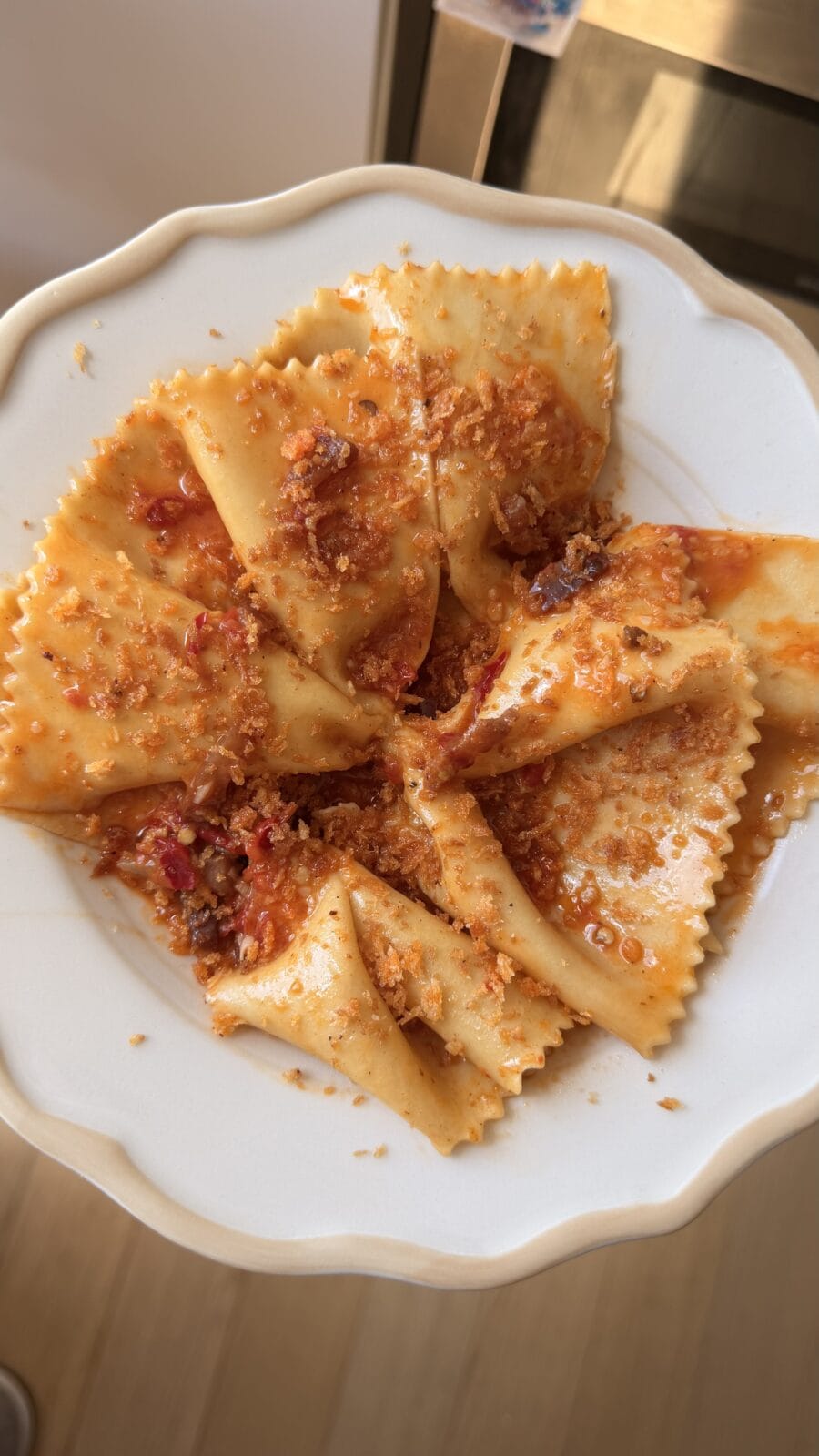 A white plate with wide, square pasta pieces topped with a chunky red sauce and sprinkled with grated cheese or breadcrumbs.