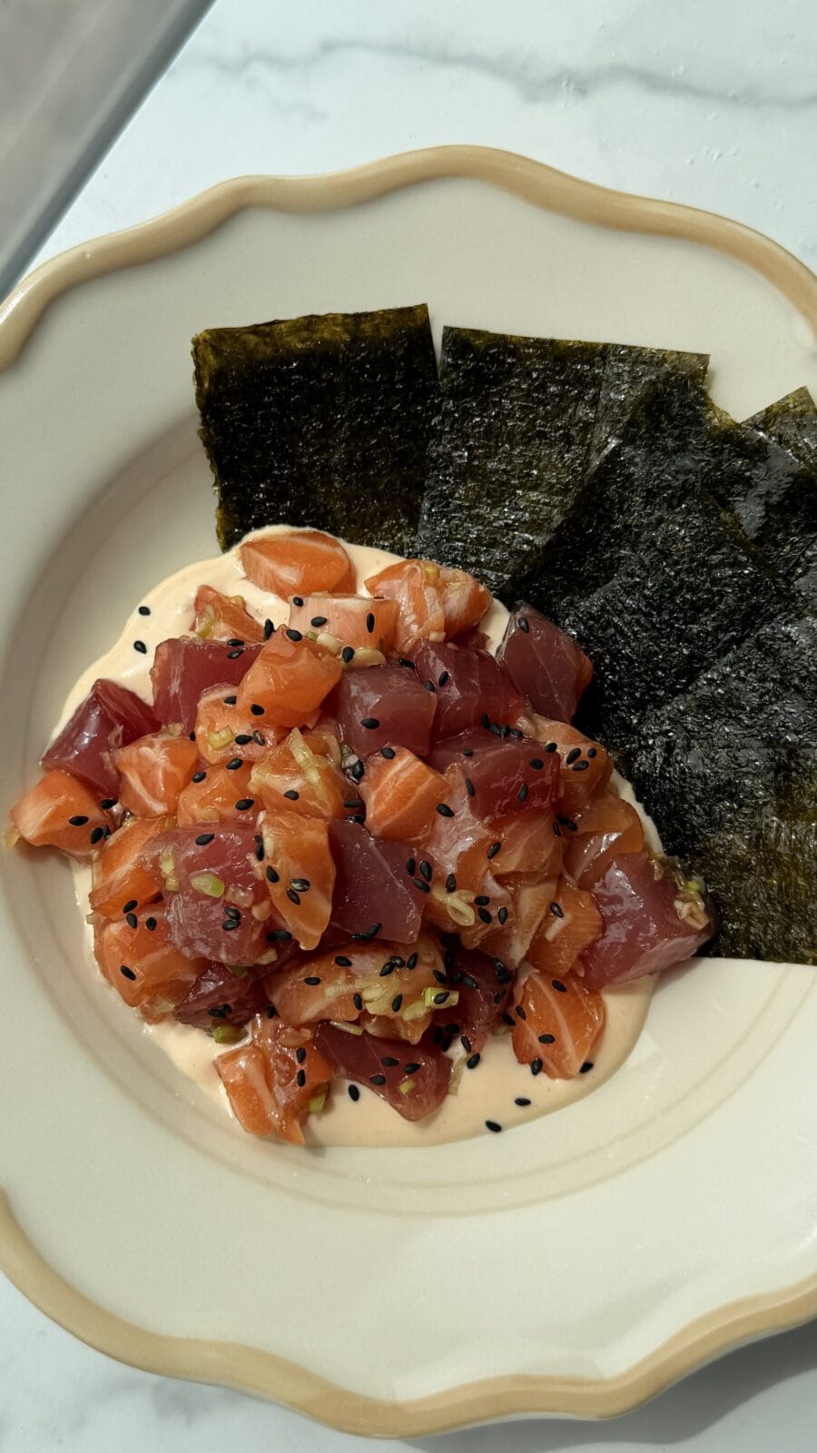 A plate with diced raw salmon and tuna on a creamy sauce, topped with black sesame seeds, served with sheets of seaweed on the side.