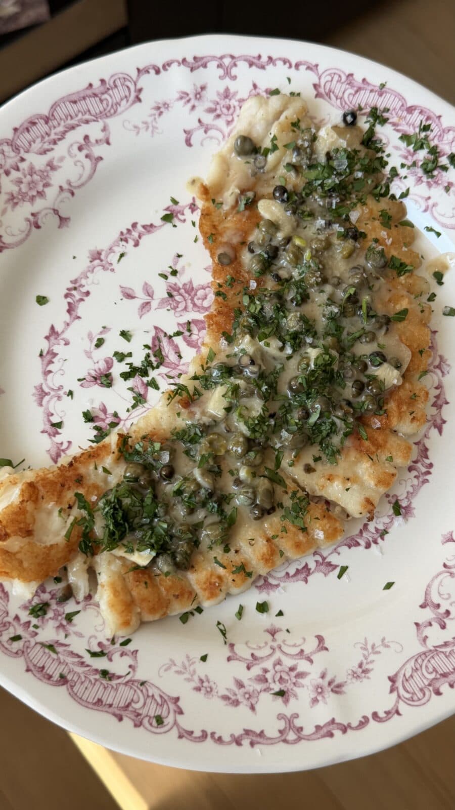A browned, crescent-shaped piece of fish topped with capers and chopped herbs on a white and pink decorative plate.