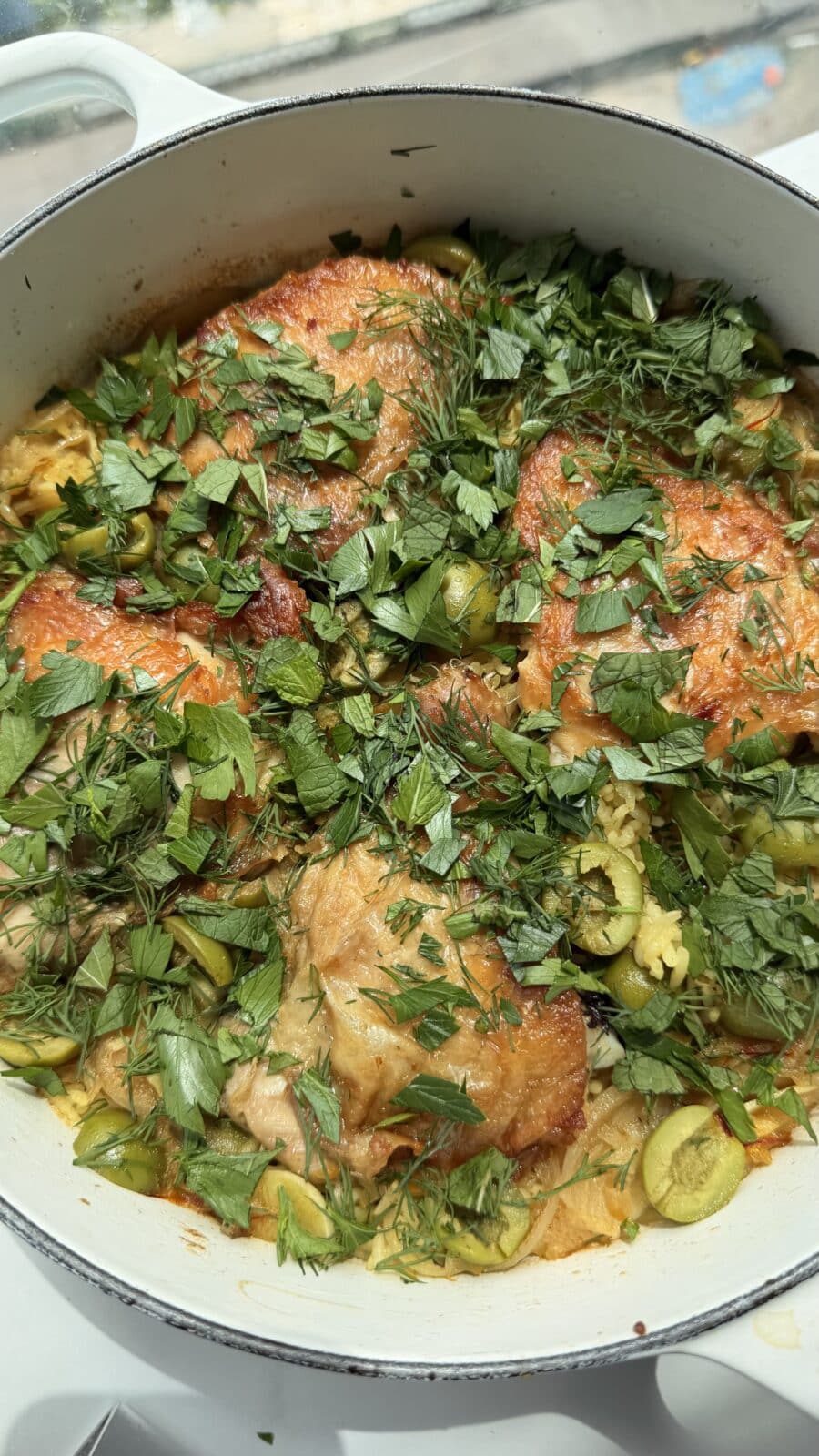 A pot of cooked chicken thighs topped with chopped fresh herbs and sliced green olives.