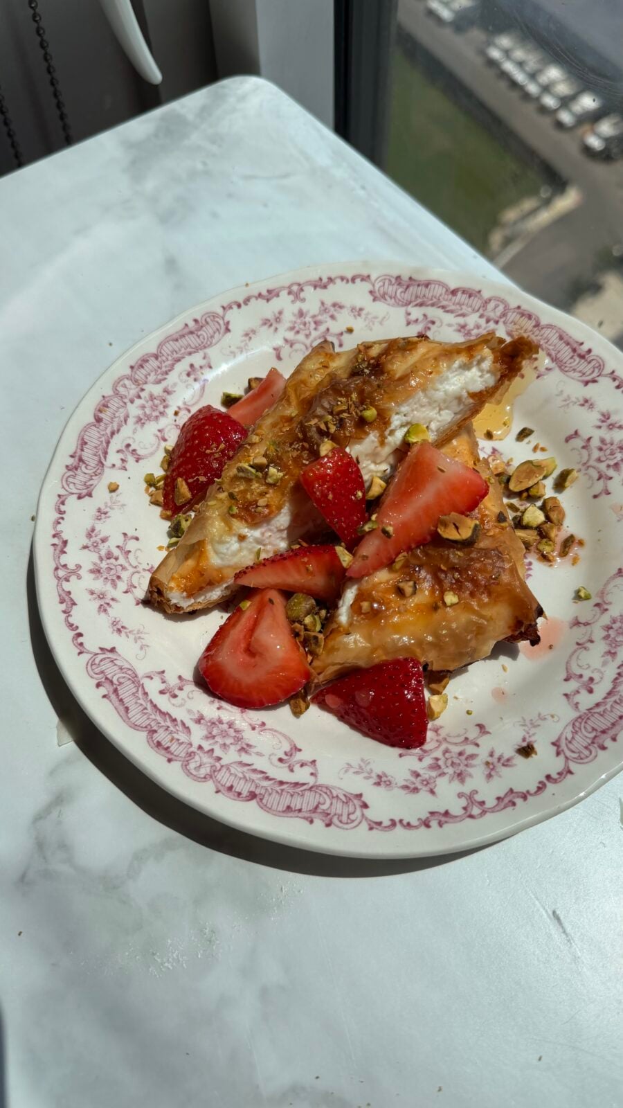 A pastry topped with fresh strawberries and chopped pistachios is served on a decorative plate, placed on a white surface near a window.