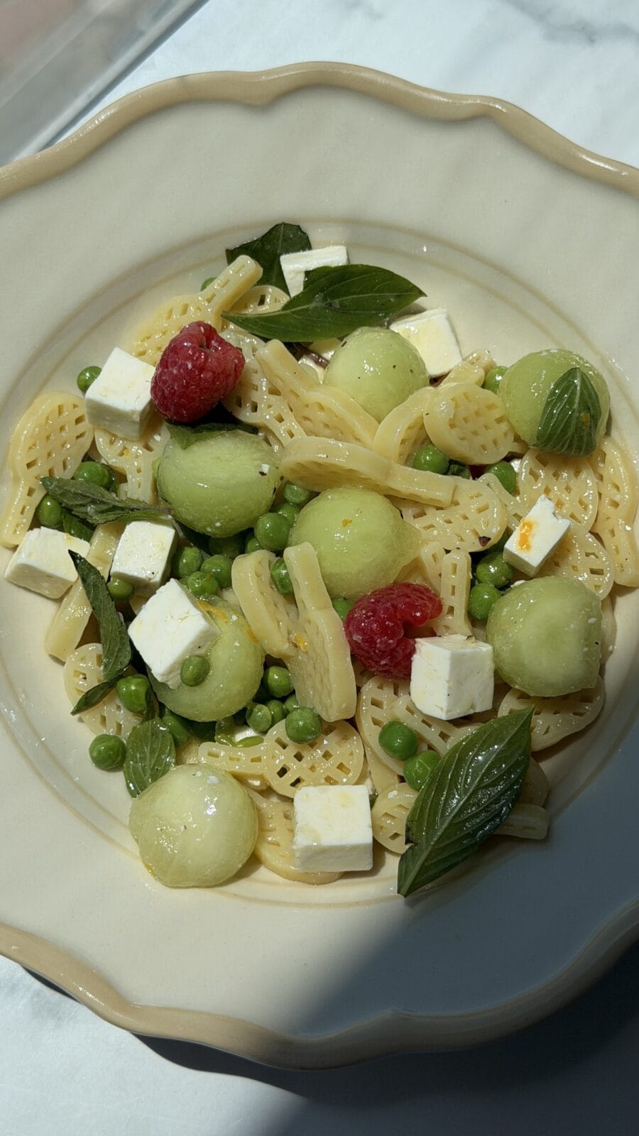 A bowl of wheel-shaped pasta with green peas, melon balls, feta cheese cubes, fresh basil leaves, and raspberries on a cream-colored plate.