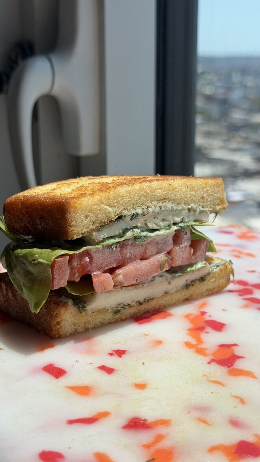 A close-up of a sandwich with lettuce, tomato, and herb spread on toasted bread, placed on a cutting board near a window with a city view in the background.