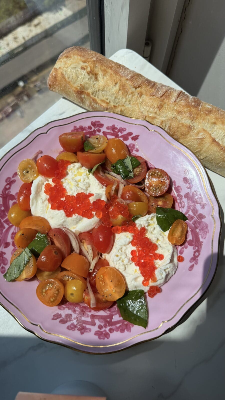 A plate of burrata cheese with cherry tomatoes, red roe, basil, and onions, served with a baguette, placed by a window in sunlight.