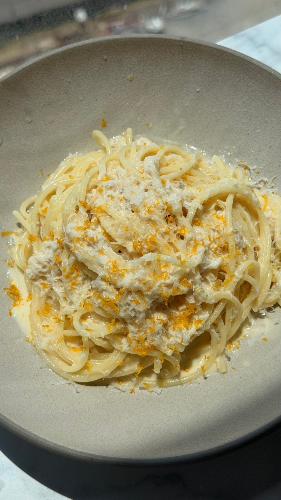 A bowl of spaghetti topped with a creamy white sauce, shredded cheese, and orange zest on a light-colored plate.