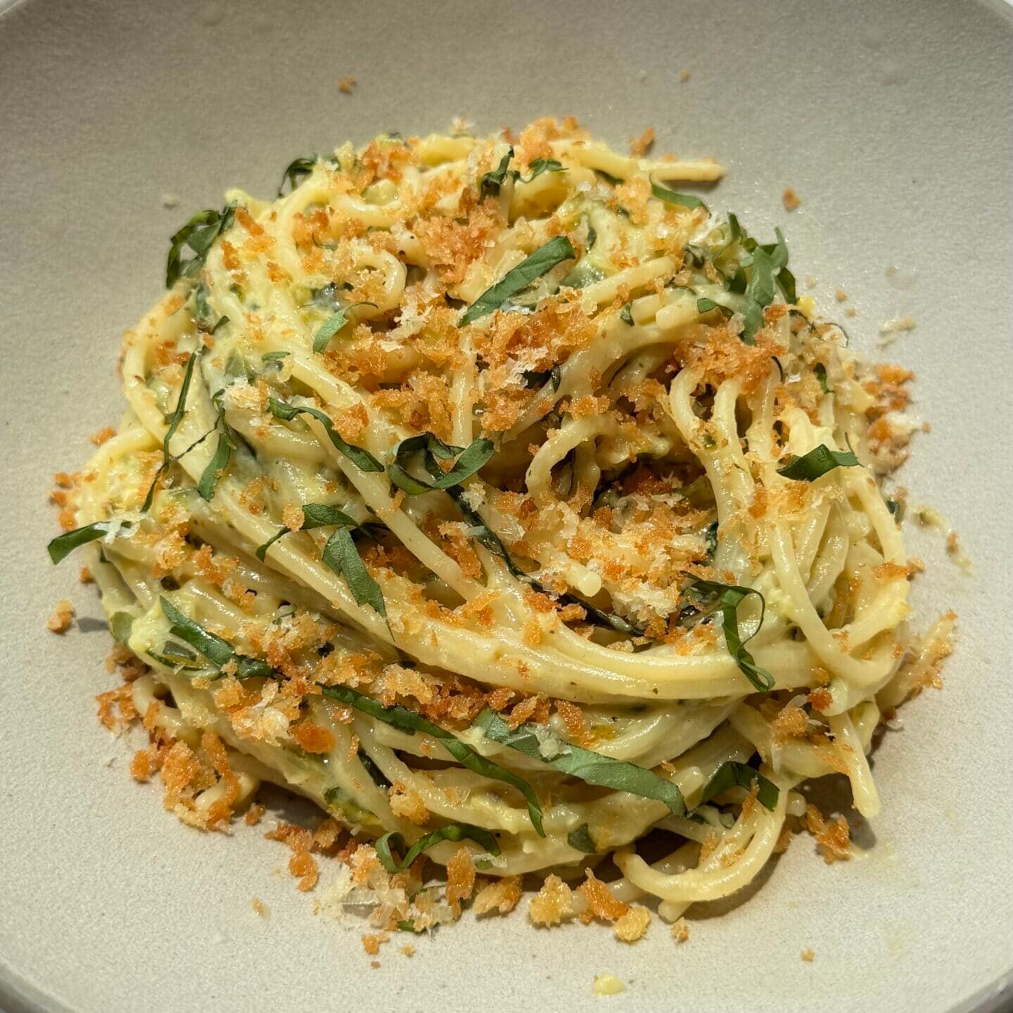 A bowl of spaghetti pasta topped with shredded greens and crispy breadcrumbs, served in a neutral-colored bowl on a light surface.