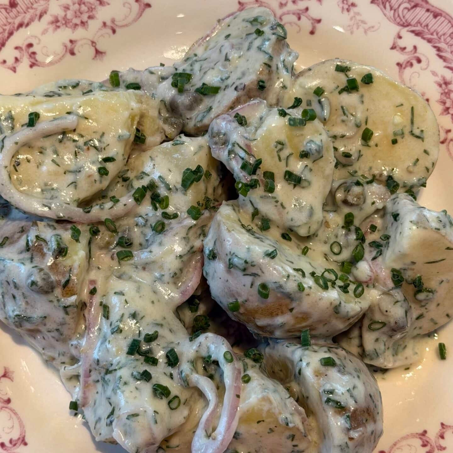 Potato salad with creamy herb dressing and sliced onions, served on a decorative plate with a pink floral border, on a gray countertop.