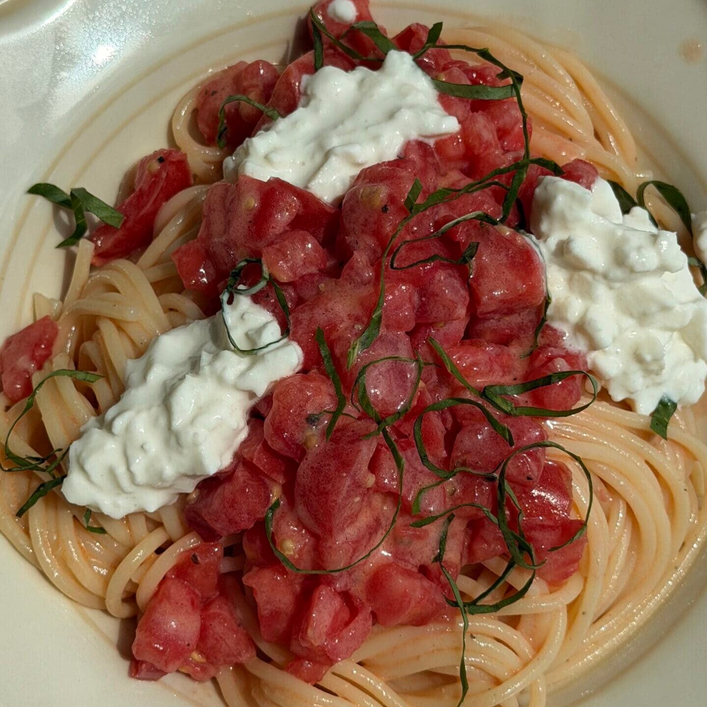 A plate of spaghetti topped with chopped tomatoes, fresh basil, and dollops of ricotta or burrata cheese on a white dish.