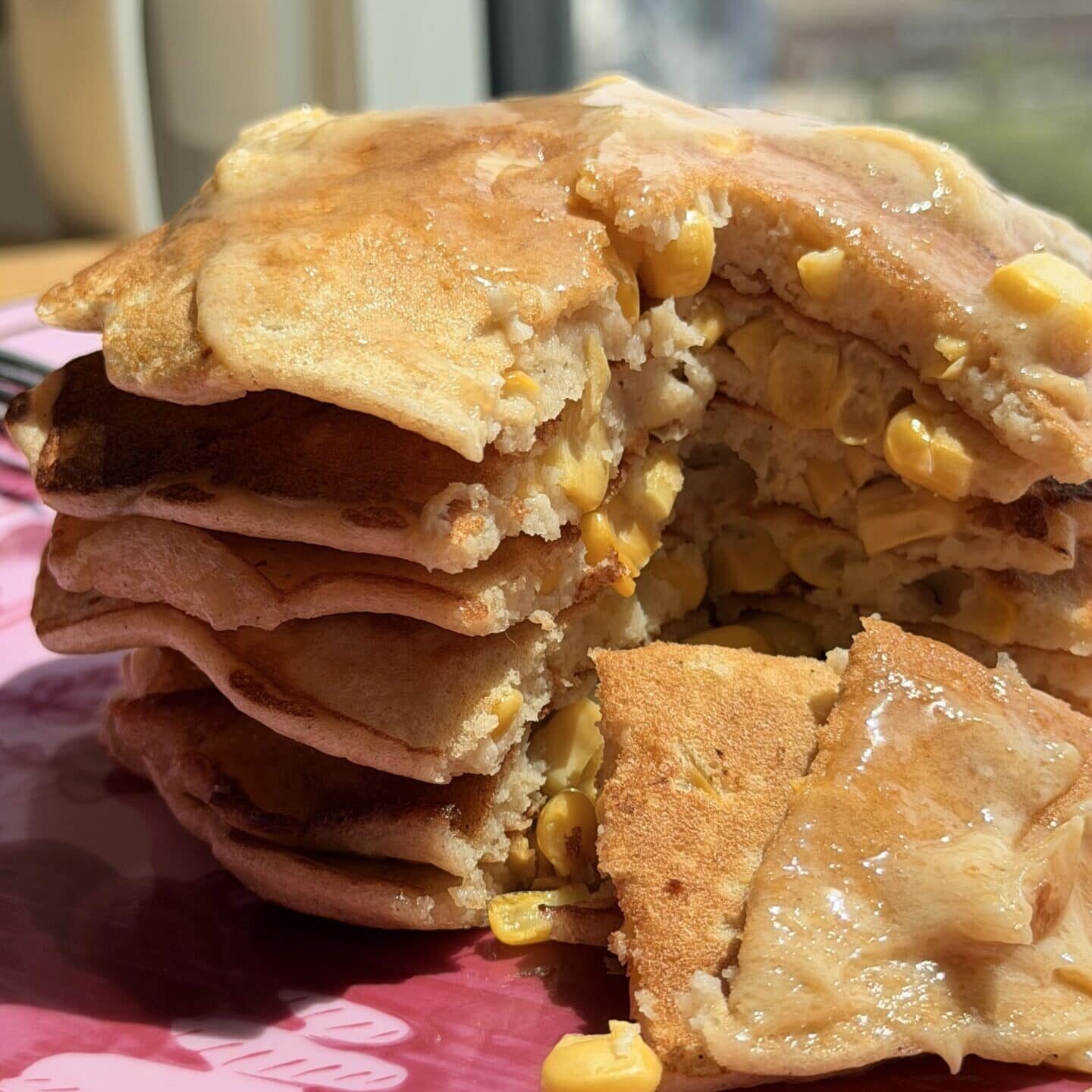 A stack of pancakes with corn kernels inside, cut to show the filling, sits on a pink plate by a window in natural light.