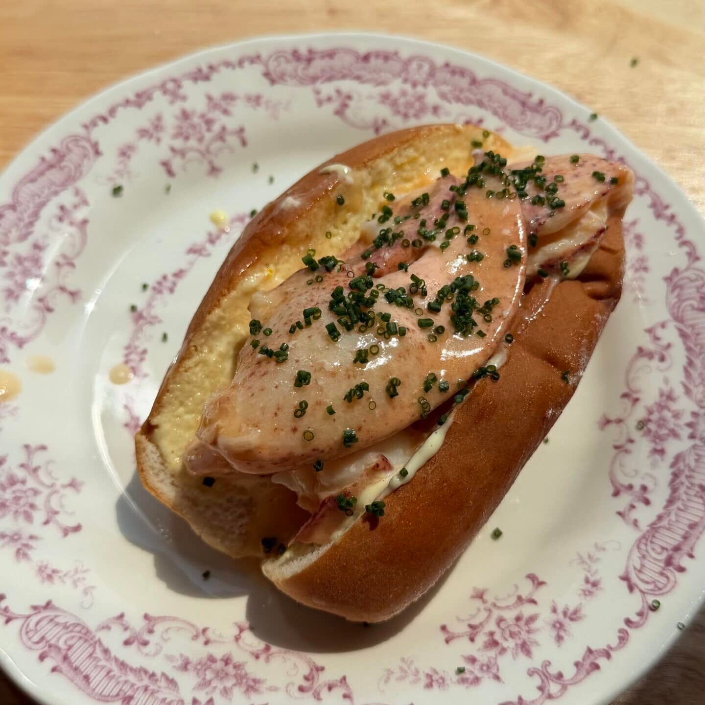 A lobster roll in a toasted bun topped with sauce and chopped herbs, served on a decorative white plate with a pink floral border.