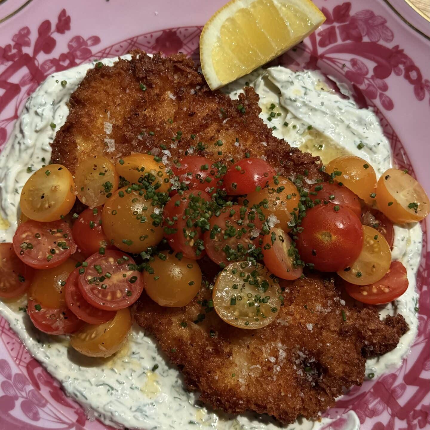 Breaded cutlet on creamy herb sauce, topped with halved cherry tomatoes and chopped chives, served with a lemon wedge on a pink floral plate.