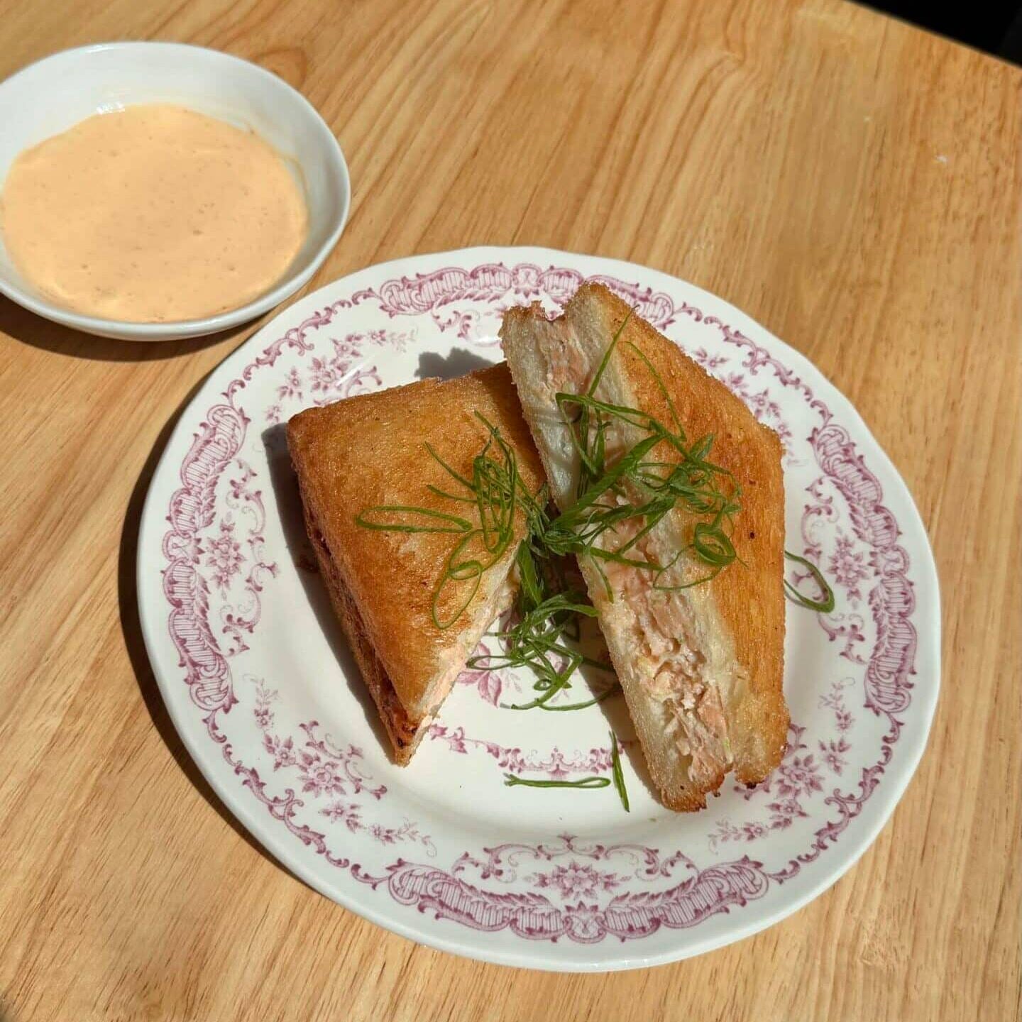 A toasted sandwich cut in half with green garnish on a patterned plate, served with a small bowl of dipping sauce on a wooden table.