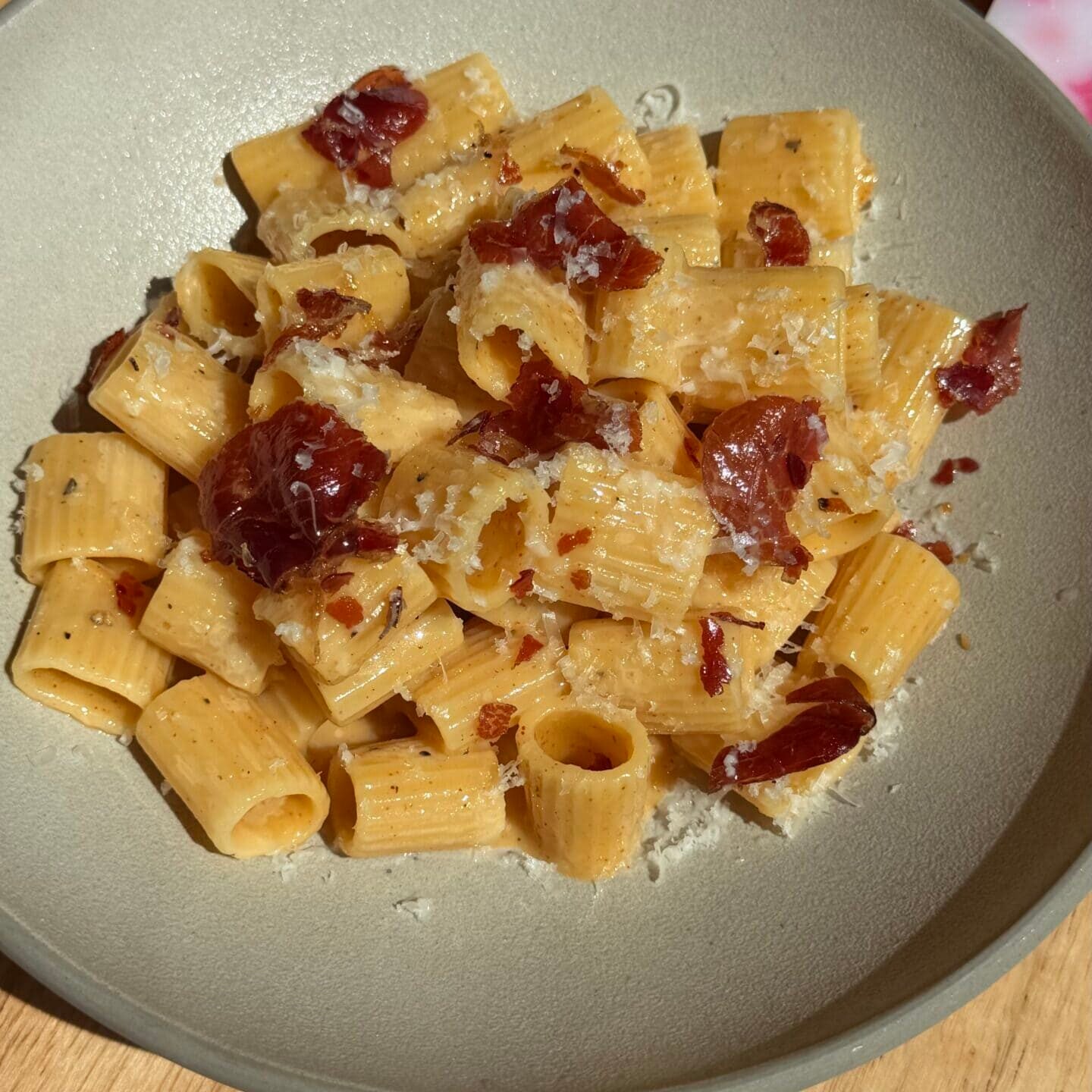 A plate of rigatoni pasta topped with grated cheese and pieces of cured meat, served on a light-colored dish on a wooden table.