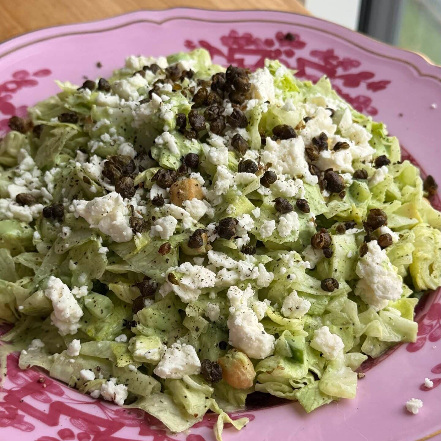 A plate of chopped lettuce salad topped with crumbled cheese, black lentils, and chickpeas, served on a pink floral dish by a window.