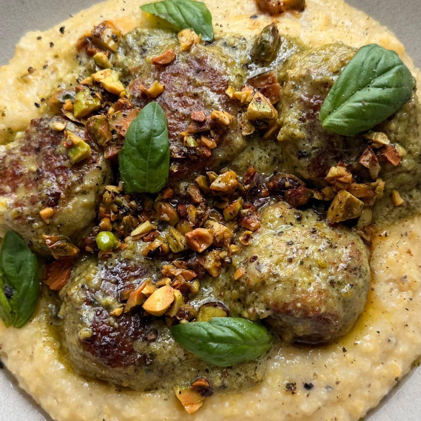 Meatballs on creamy polenta, topped with pesto, chopped pistachios, black pepper, and fresh basil leaves, served in a beige bowl.