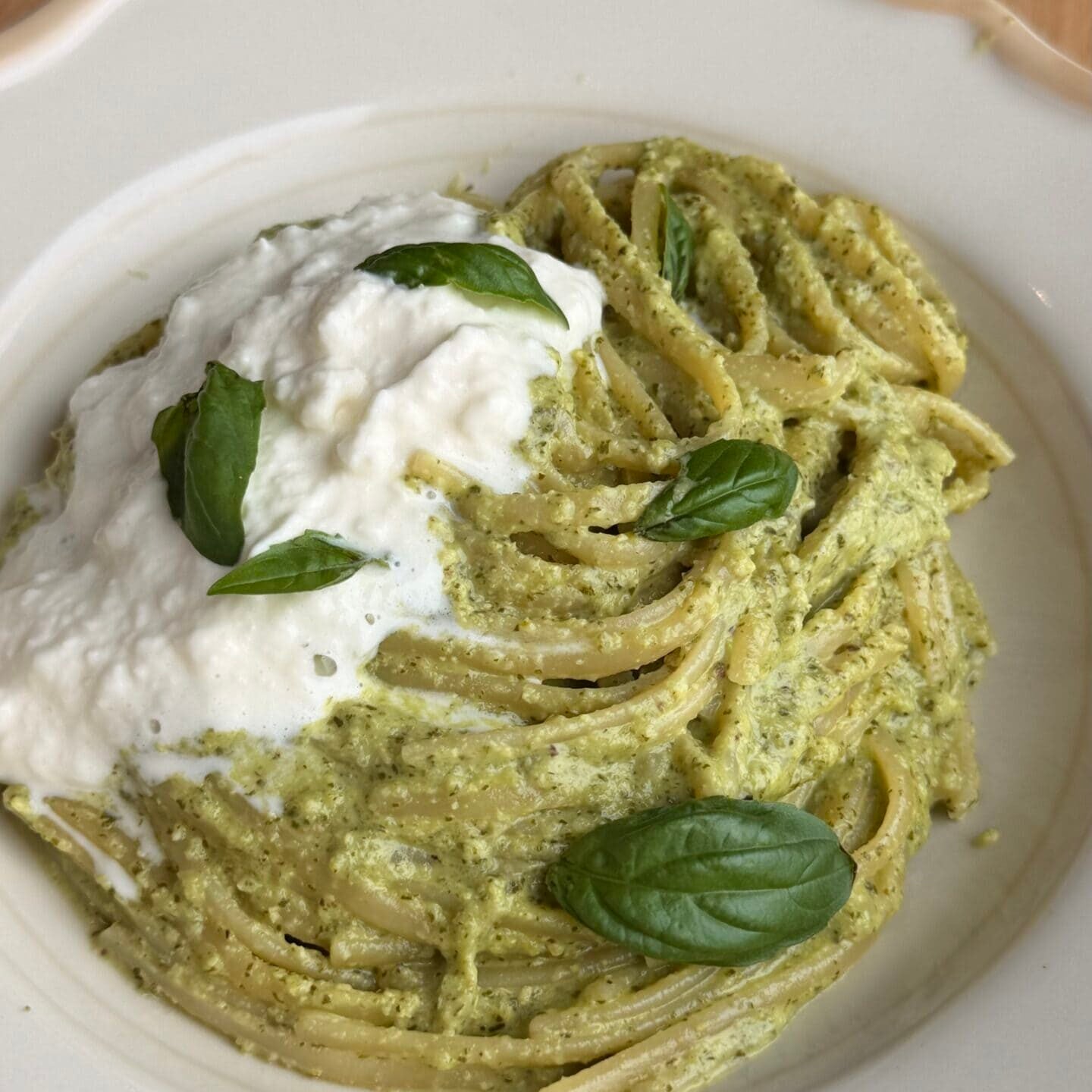 A plate of pasta with green pesto sauce, topped with a dollop of white cheese and garnished with fresh basil leaves on a wooden table.