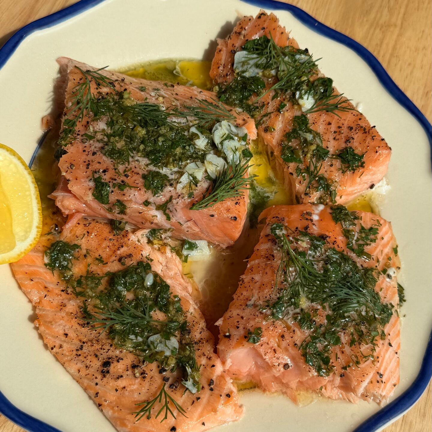 Four salmon fillets seasoned with herbs and garlic on a plate with a lemon wedge.