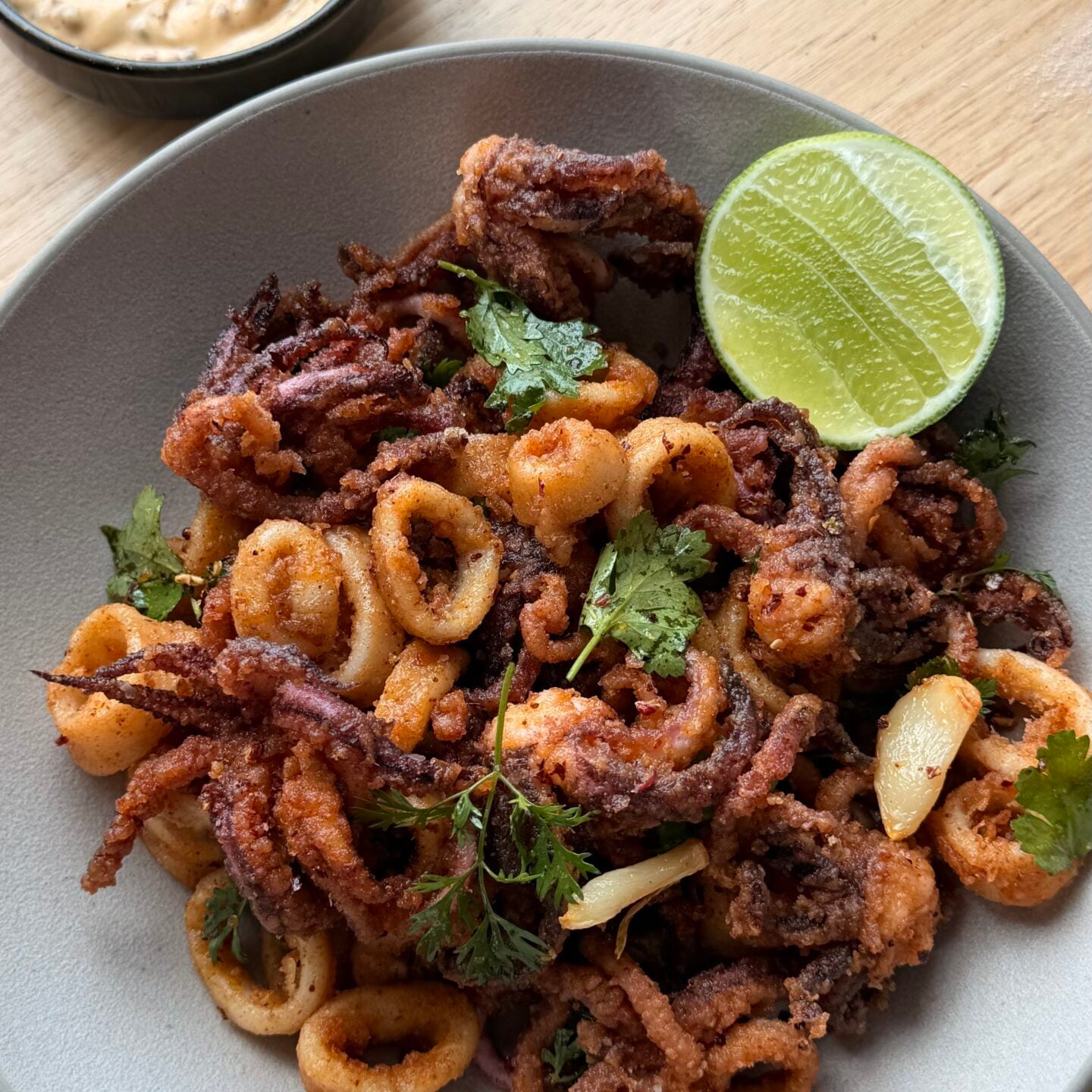 A bowl of fried calamari garnished with cilantro and a lime wedge, served with a small dish of creamy dipping sauce on a wooden surface.
