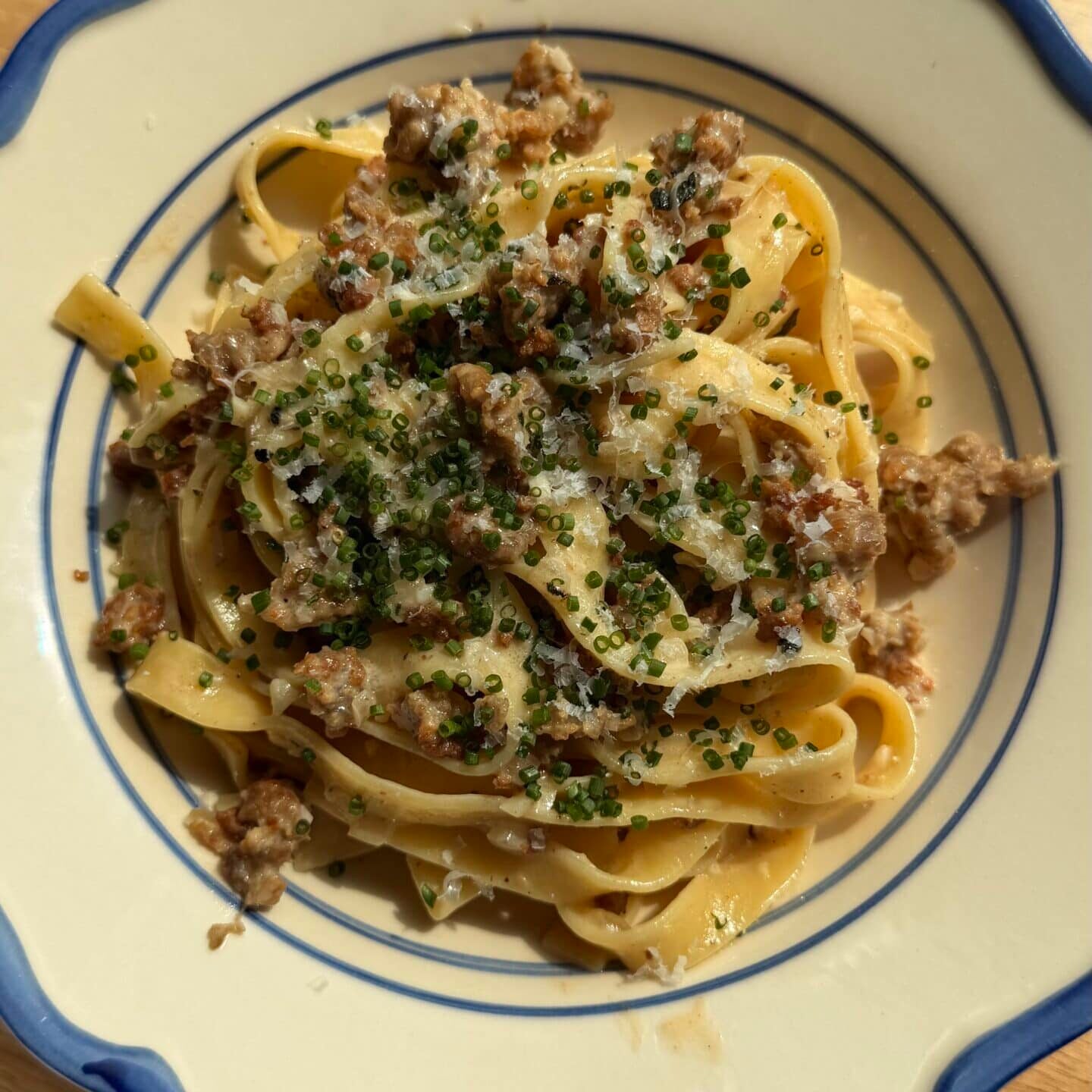 A plate of tagliatelle pasta topped with ground meat, grated cheese, and chopped herbs, served on a white dish with blue trim.