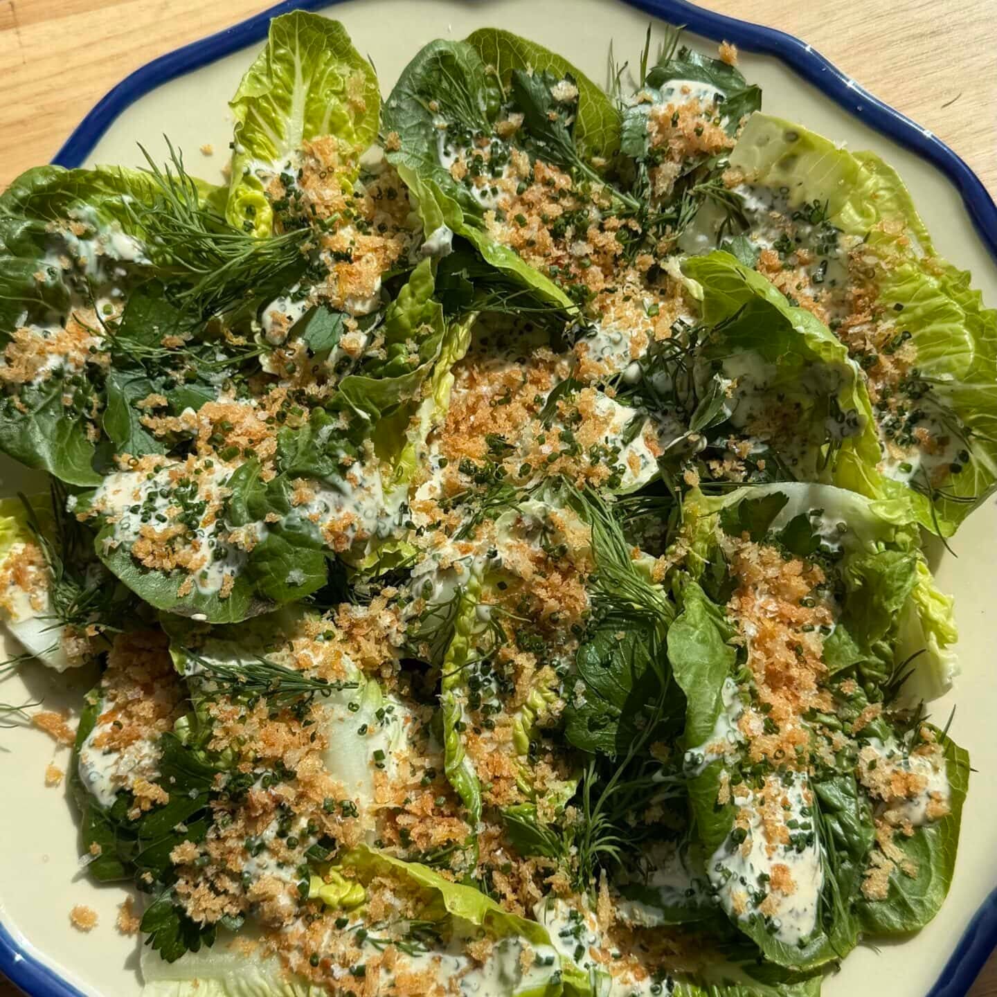 A plate of leafy green salad topped with breadcrumbs, fresh herbs, and a creamy dressing, placed on a wooden surface in sunlight.